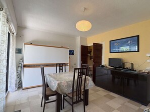 A dining area with a table, chairs, a whiteboard, a television, and a kitchen area with cabinets.