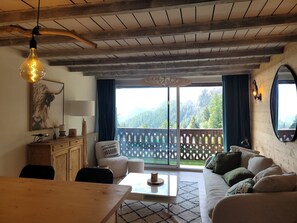 Uma sala de estar acolhedora com teto de madeira, um sofá, uma mesa de centro e vista para uma paisagem montanhosa através de grandes janelas.