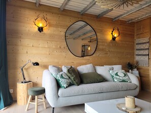 Ein gemütliches Zimmer mit Holzbalkendecke, einem runden Spiegel, einem hellen Sofa mit Zierkissen, einem kleinen Holzschemel und einem Tisch mit einem gewebten Korb.