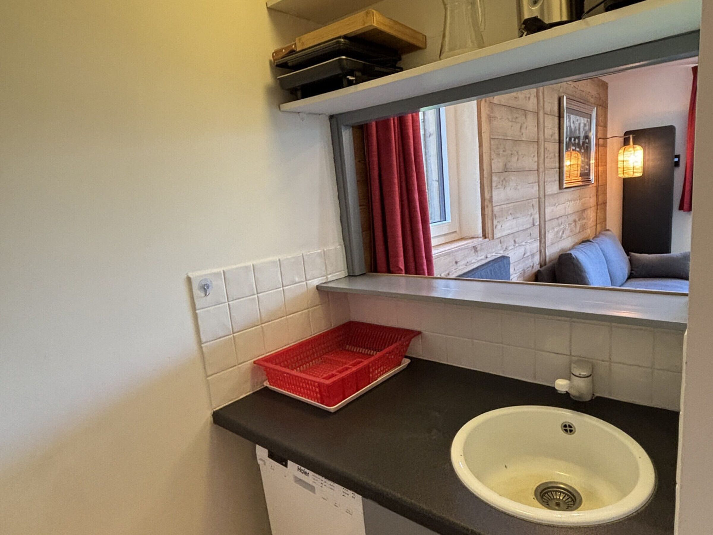 A compact bathroom with a white sink, a red basket, and a window with red curtains.
