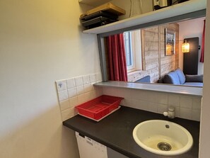 Ein kleines Badezimmer mit einem weißen Waschbecken, einem roten Korb und einem Fenster mit roten Vorhängen.