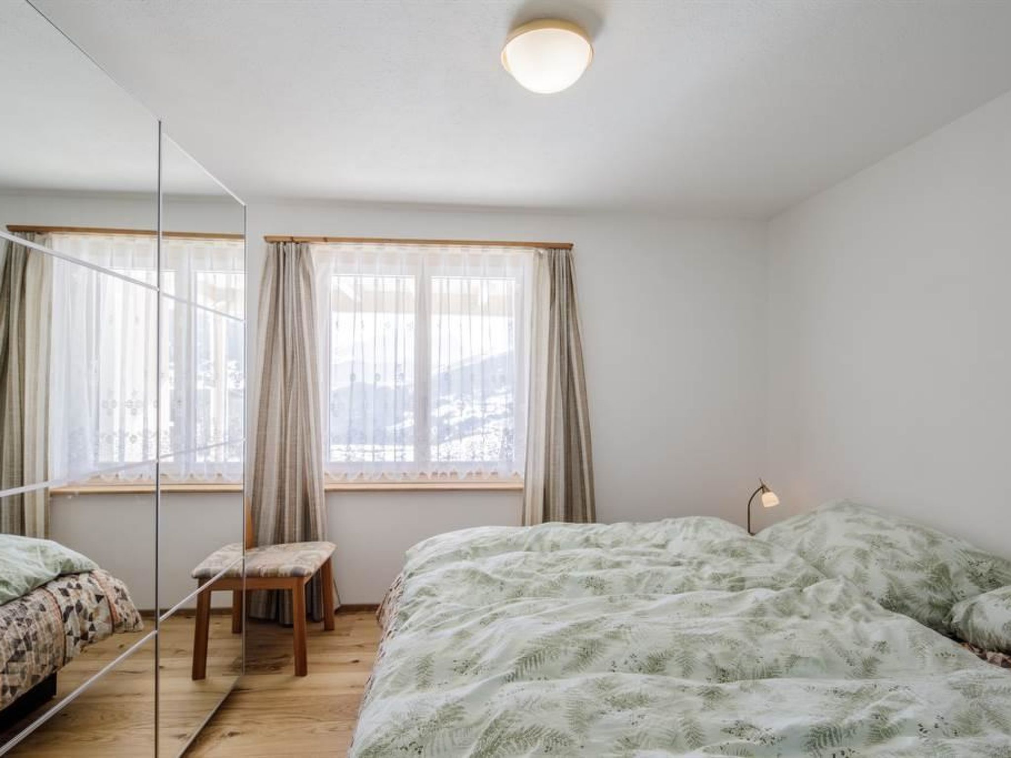 A bedroom with two beds, a chair, a bedside table, and a window with curtains.