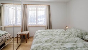 A bedroom with two beds, a chair, a bedside table, and a window with curtains. - Guarda Val Müller Erlenstube Wohnung Erlenstube Wohnung Erlenstube by Interhome (Lantsch)