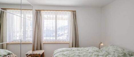 Ein Schlafzimmer mit zwei Betten, einem Sessel, einem Nachttisch und einem Fenster mit Vorhängen.