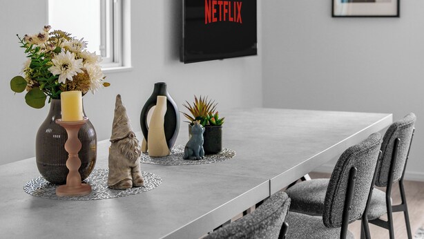 Un espace repas moderne avec une longue table, des chaises, un vase de fleurs, une bougie et un téléviseur fixé au mur qui affiche le logo de Netflix.