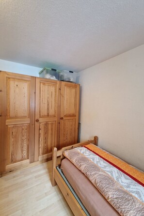 A bedroom with a wooden wardrobe, a bed with a patterned bedspread, and a small storage unit on the wardrobe.