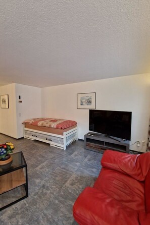 Ein Wohnzimmer mit Bett, einem roten Sofa, einem Fernseher und einem kleinen Tisch mit Blumen.