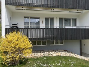 A modern two-story building with a balcony, a small garden area with rocks and a yellow flowering tree, and a clear sky. - Tga Crap Alv Appartement/Fewo, Dusche, WC, 2 Schlafräume by Interhome (Savognin)