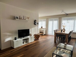 A living room with a flat-screen TV, a dining area with a wooden table and chairs, and a large window with sheer curtains. - Tga Crap Alv Appartement/Fewo, Dusche, WC, 2 Schlafräume by Interhome (Savognin)