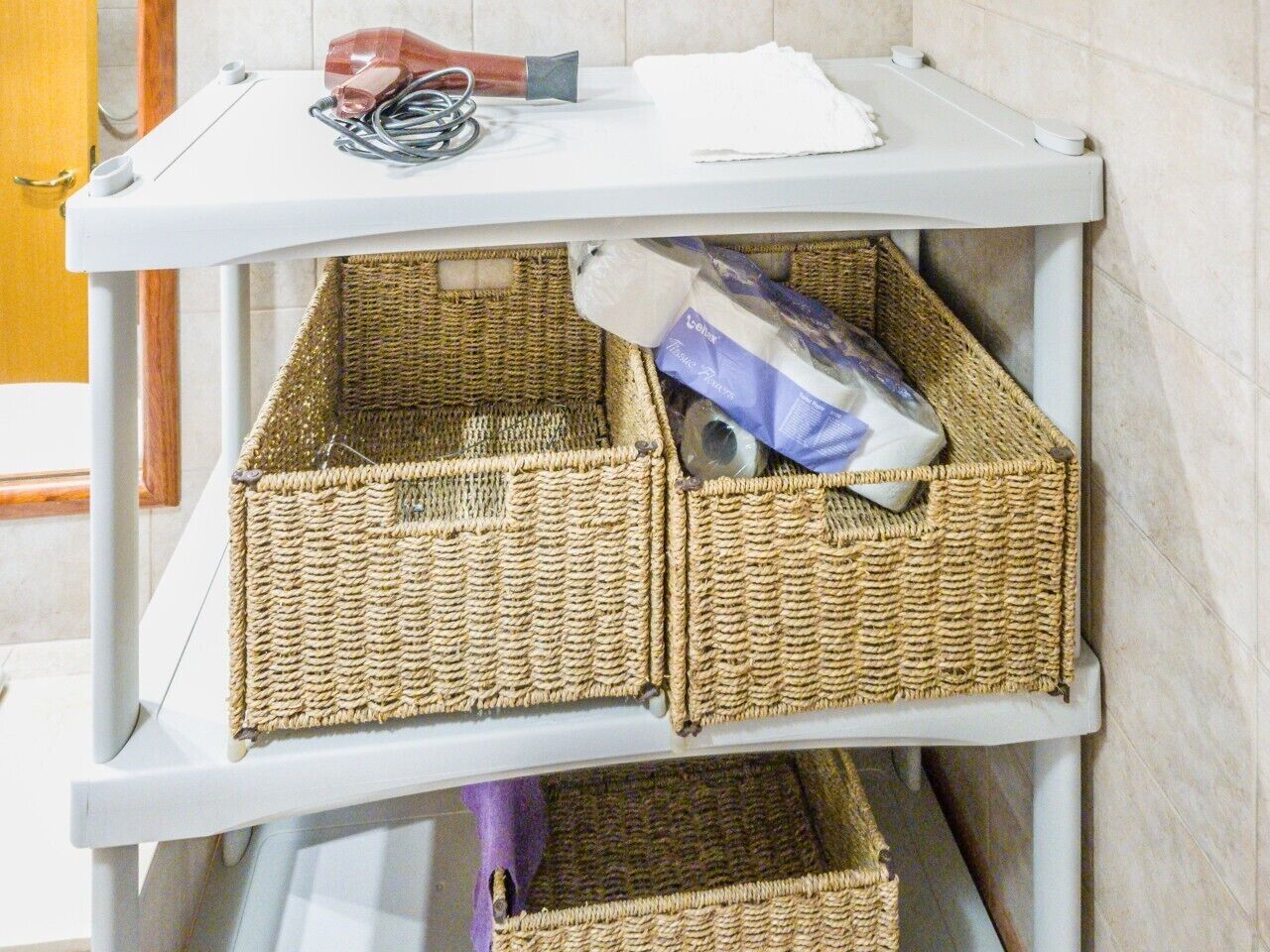 A white metal cart with wicker baskets containing items like a bag of cat litter and a roll of toilet paper.