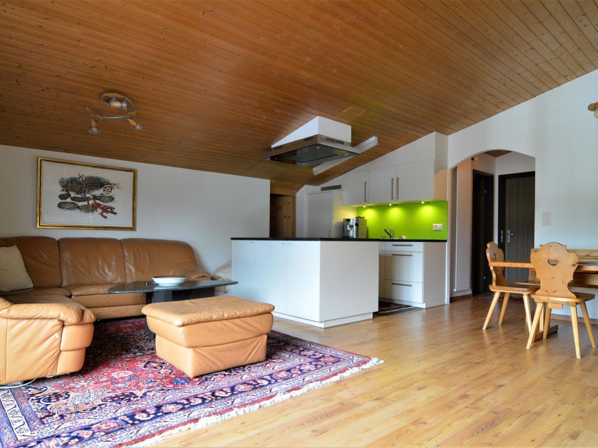 A modern living space with a wooden ceiling, a sofa set, a dining area with wooden chairs, and a kitchen with a green accent wall.