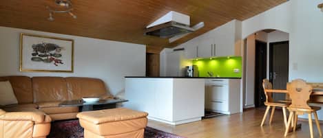 A modern living space with a wooden ceiling, a sofa set, a dining area with wooden chairs, and a kitchen with a green accent wall.