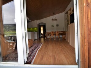 Een open woonkamer met een eettafel en stoelen, uitzicht op de omgeving en een keuken op de achtergrond.