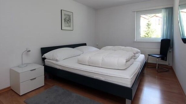Ein Schlafzimmer mit Bett, Nachttisch, Stuhl, Fenster und einem Bild an der Wand.