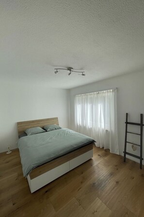 Ein Schlafzimmer mit einem hölzernen Bett, weißen Wänden, einer Deckenleuchte und einem Fenster mit durchsichtigen Vorhängen.