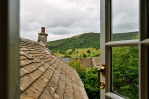 Blick durch ein Fenster auf ein Gebäude mit traditionellem Stein-Dach und Schornstein, mit Blick auf ein grünes Tal mit sanften Hügeln und vereinzelten Bäumen.