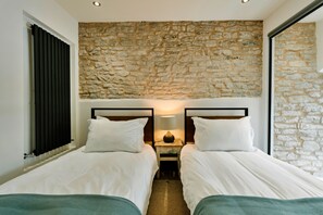 A bedroom with two beds, a stone wall, a nightstand with a lamp, and a window with blinds. - Heather View Chapel (Bradwell)