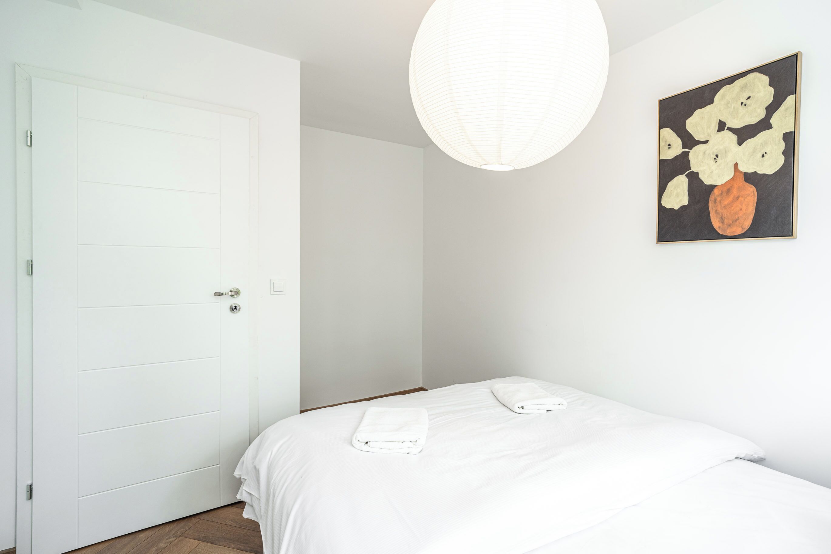 Un dormitorio con una puerta blanca, una cama con ropa de cama blanca y una lámpara fijada a la pared.