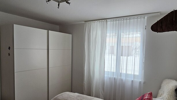 Um quarto com uma cama, um guarda-roupa e uma janela com cortinas translúcidas.