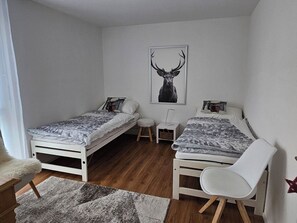 A small, well-lit bedroom with two single beds, a white chair, a small table, and a framed deer picture on the wall.