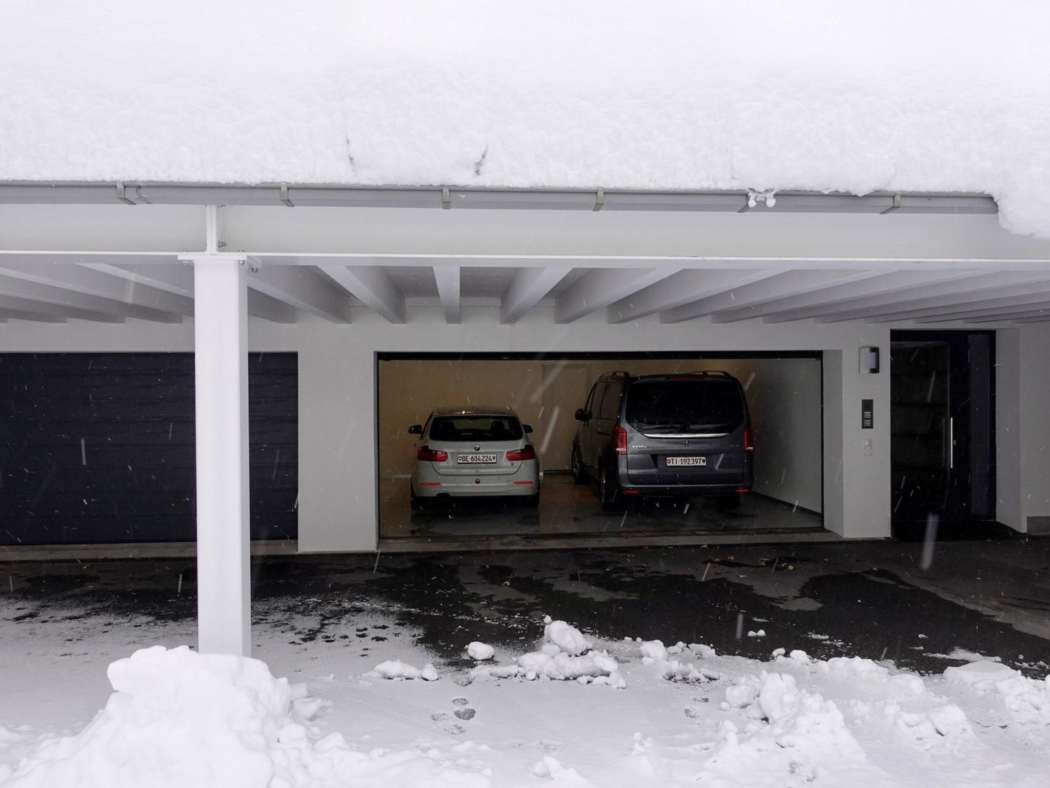Un parcheggio coperto con due automobili al suo interno, neve a terra e una porta parzialmente visibile a destra.