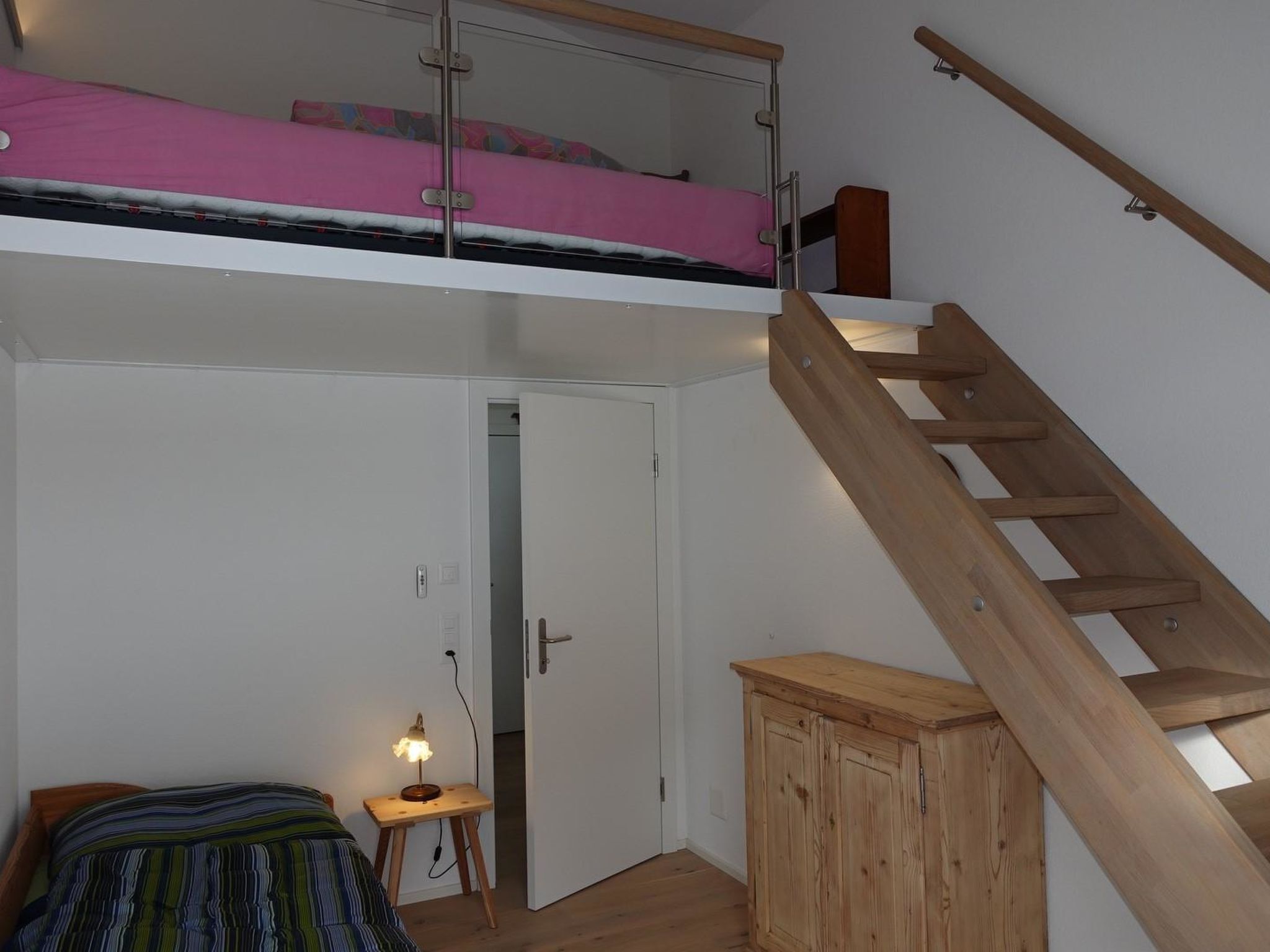 A room with a bunk bed, a wooden ladder, a desk with a lamp, and a wardrobe.