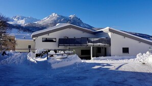 Un edificio moderno de dos pisos con techo plano y ventanas amplias, rodeado de nieve y montañas al fondo.