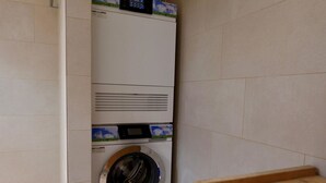 Una lavadora y secadora compactas, fijadas a la pared, en un pequeño lavadero con paredes de azulejos y un banco de madera.