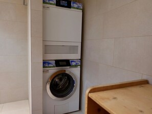 A compact, wall-mounted washer and dryer unit in a small laundry area with tiled walls and a wooden bench. - Casa Stella Appartement/Fewo, Dusche, WC, 2 Schlafräume by Interhome (Savognin)