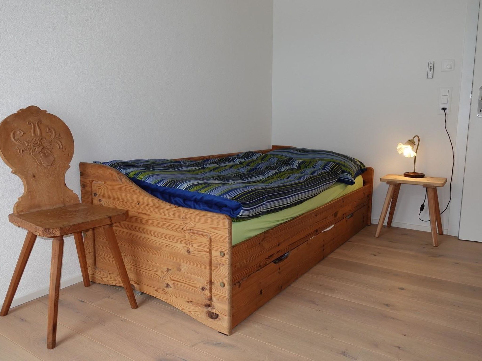 A single bed with a wooden frame, a small bedside table with a lamp, and a chair with a carved backrest.