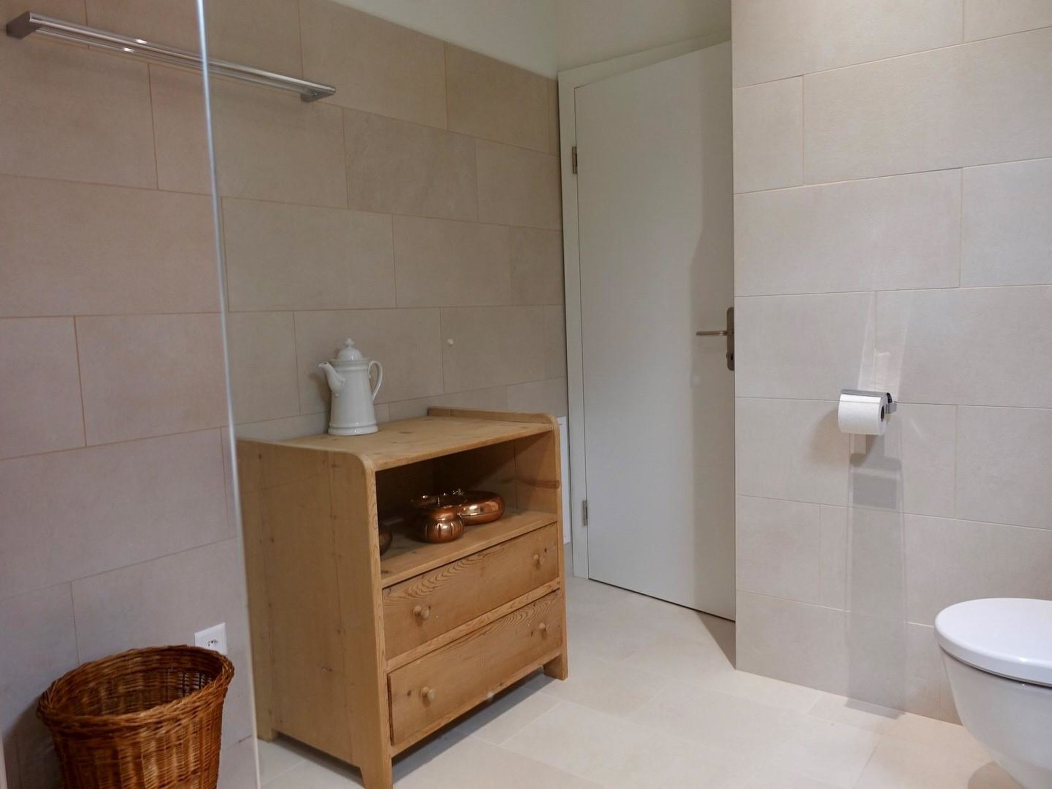 Een badkamer met toilet, een houten kast met lades, een rieten mand en een douche met glazen deur.