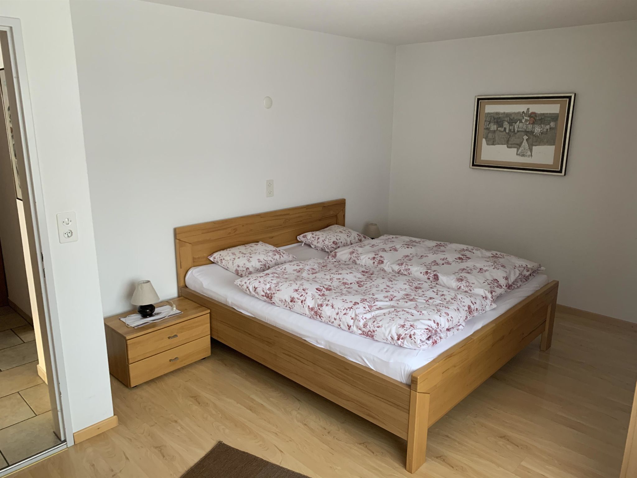 Une chambre avec un lit simple, une tête de lit en bois, une table de chevet avec une lampe et un tableau encadré au mur.