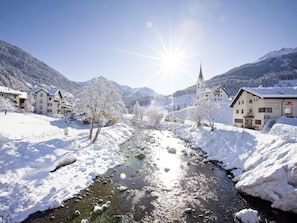 Ein verschneites Dorf mit einem Fluss, Gebäuden und Bergen im Hintergrund.