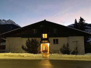 Um chalé de dois andares com uma entrada iluminada e acolhedora, rodeado por neve e montanhas.