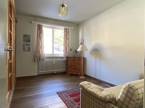 Ein Zimmer mit Holzboden, Heizkörper, einem fenster mit Vorhängen, Holzschrank, Lampe und einem karierten Sofa.