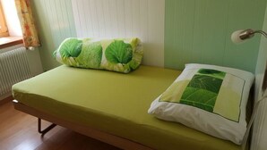Uma cama de solteiro com colchão verde e duas almofadas, uma com padrão de folha e outra lisa, branca.