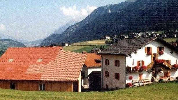Paisagem rural com um celeiro e um edifício branco com telhado vermelho, rodeado por campos verdes e montanhas ao fundo.