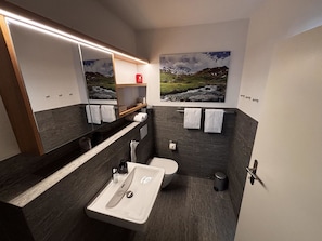 A modern bathroom with a sink, toilet, and shower. There are towels and a red emergency kit on the wall. - Bergnest 4 Appartement/Fewo, Bad, WC, 2 Schlafräume by Interhome (Bivio)