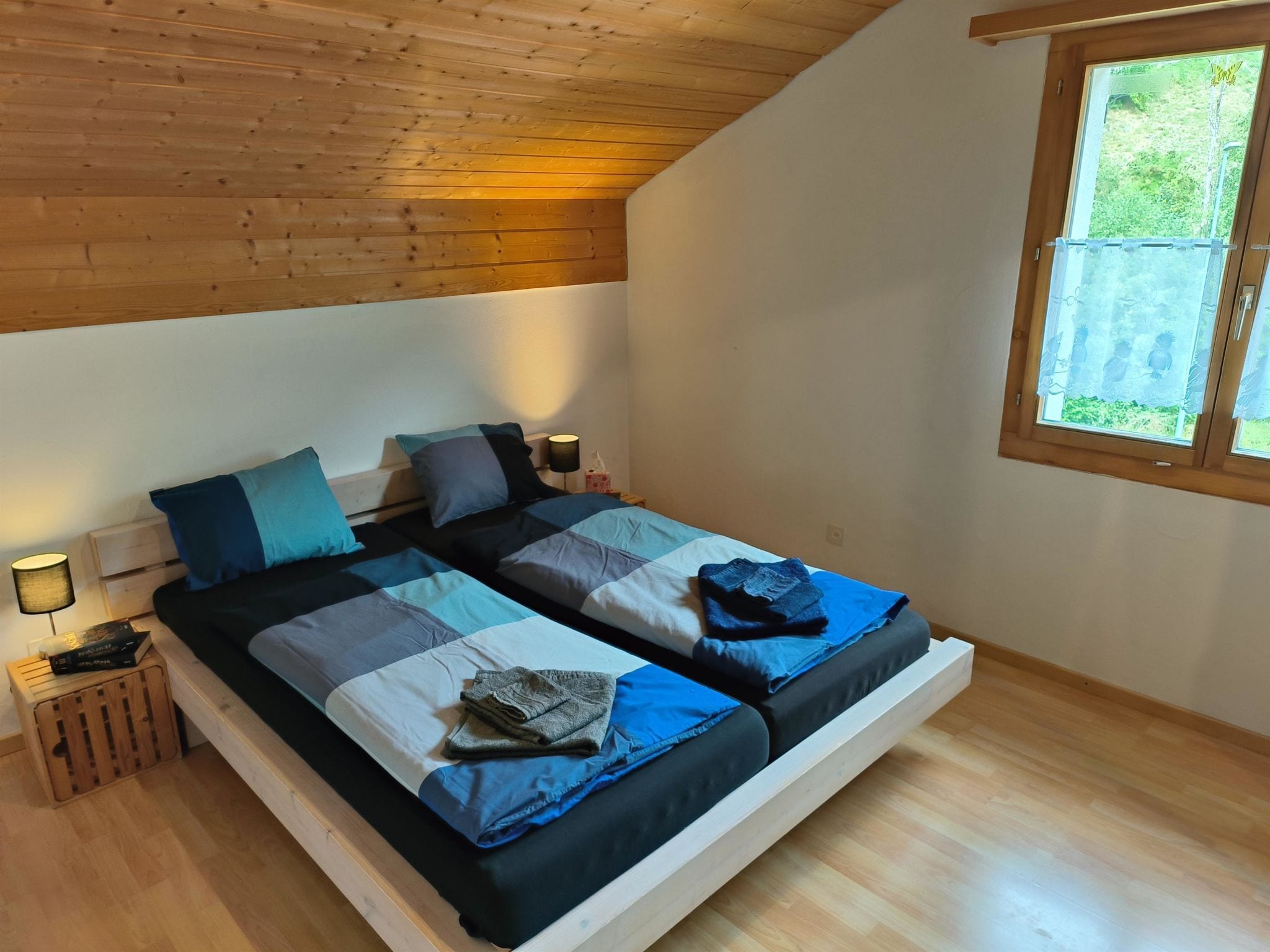 Ein Schlafzimmer mit Holzbalkendecke, einem Bett mit blau-grauem Bettzeug, einem Fenster mit Blick auf Grünflächen und einem Nachttisch mit Lampe.