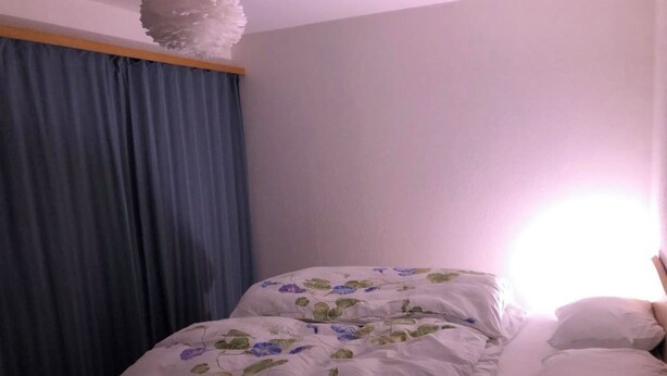 Quarto com cama, janela com cortinas azuis e luminária fixada na parede.