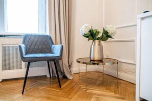 A room with a blue upholstered chair, a round table with a metallic base, and a vase with white flowers. - Unbeatable Location at March 15 Sq: Quiet, Elegant & Scenic – 333 (Budapest)