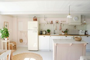 A kitchen with white cabinetry, a refrigerator, a sink, and a dining area with a table and chairs. - Thatched idyll from the 1800s in Voldtofte (Glamsbjerg)