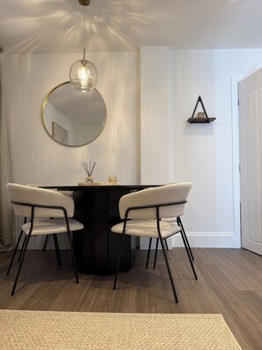 Ein Essbereich mit einem runden Tisch, zwei Stühlen, einer Hängeleuchte, einem an der Wand befestigten Spiegel und einem kleinen Regal.