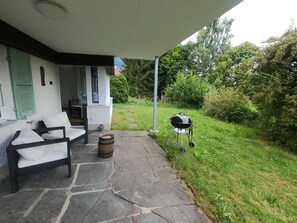 A covered patio with outdoor furniture, a barbecue grill, and a stone pathway leading to a house.