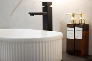 Ein Badezimmer mit einem weißen Waschbecken, einem schwarzen Wasserhahn und zwei braunen Flaschen, beschriftet mit 'HAND WASH' und 'SHOWER GEL'.