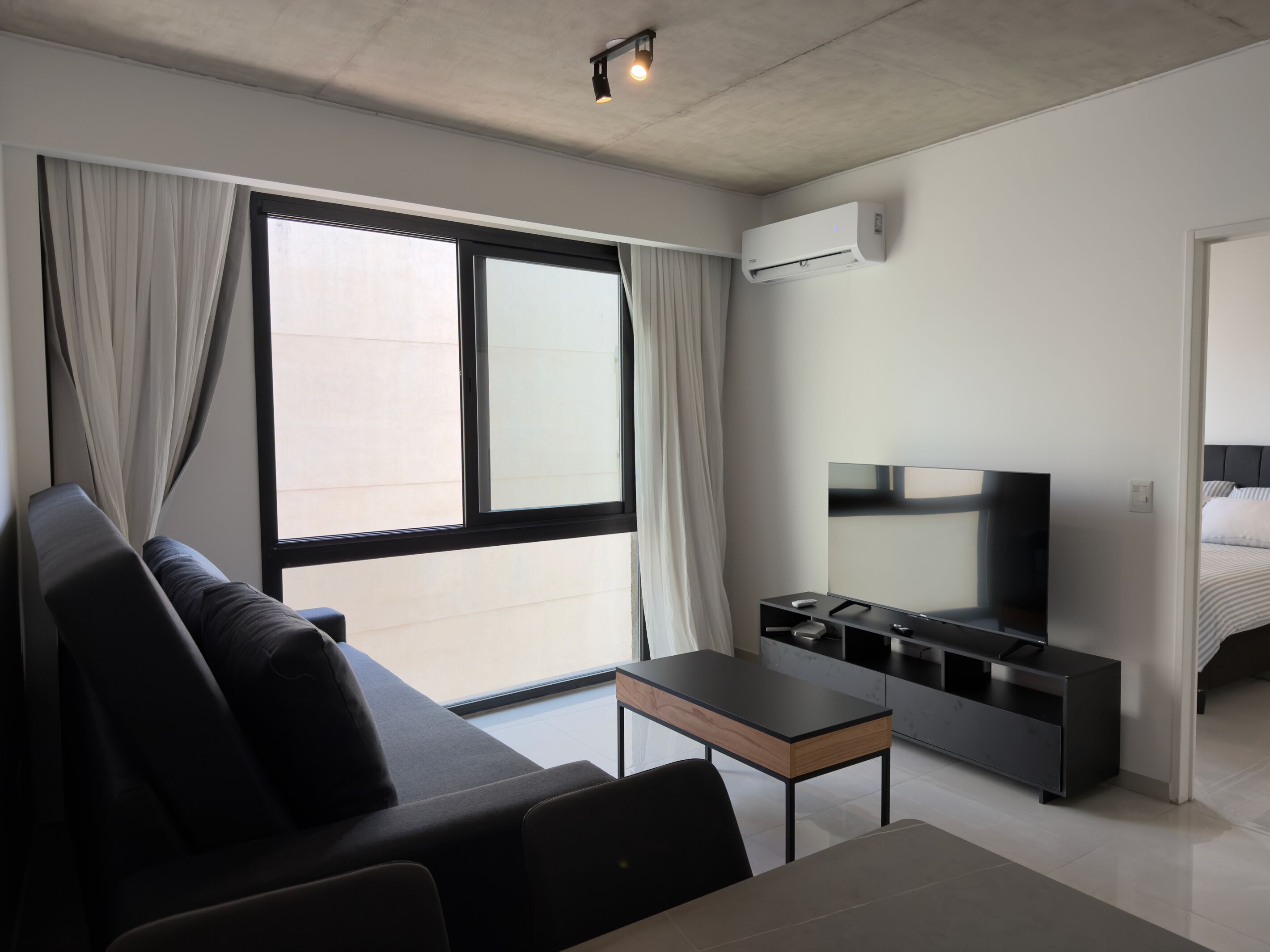 A modern hotel room with a sofa, a TV, a small table, and a large window with curtains.