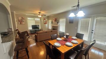A dining area with a wooden table set for four, surrounded by chairs, in a room with a ceiling fan, a television in the background, and a fireplace.