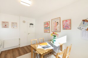 Un coin repas avec une table en bois, des chaises et un vase de fleurs. Les murs sont décorés et il y a une porte avec une horloge.