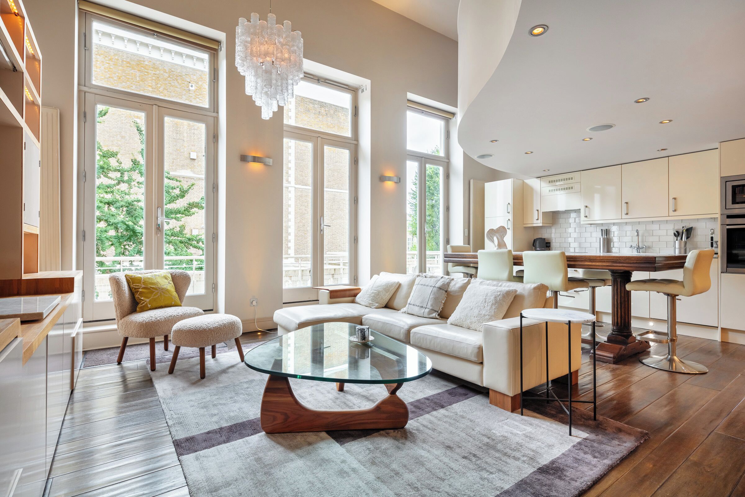 A modern living room with a glass coffee table, a sectional sofa, and a dining area with a wooden table and bar stools.