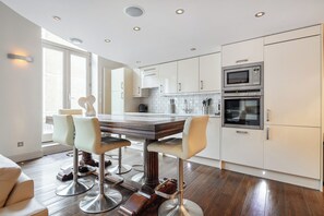 A modern kitchen with white cabinets, stainless steel appliances, and a wooden dining table with bar stools. - Elegant Earls Court Apartment | Loft Bedroom | Lift Access (London)